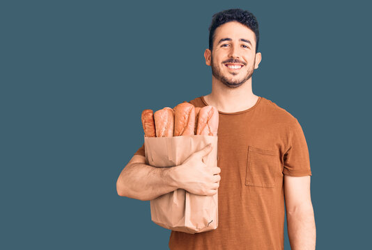 Young hispanic man holding paper bag with bread looking positive and happy standing and smiling with a confident smile showing teeth
