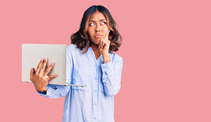 Young beautiful mixed race woman working using computer laptop serious face thinking about question with hand on chin, thoughtful about confusing idea