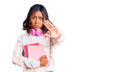 Young beautiful mixed race woman holding books wearing headphones with open hand doing stop sign with serious and confident expression, defense gesture