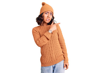 Young beautiful mixed race woman wearing wool sweater and winter hat pointing aside worried and nervous with forefinger, concerned and surprised expression