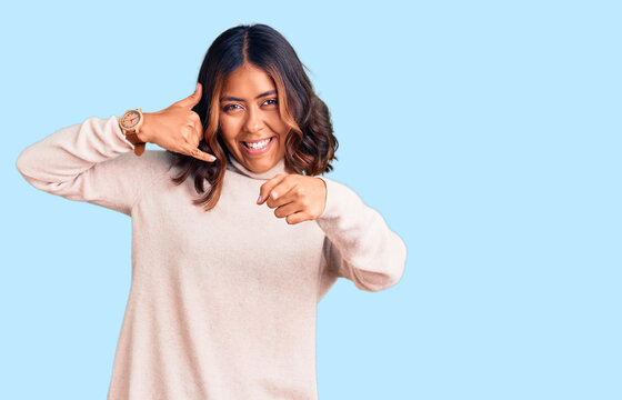 Young beautiful mixed race woman wearing winter turtleneck sweater smiling doing talking on the telephone gesture and pointing to you. call me.
