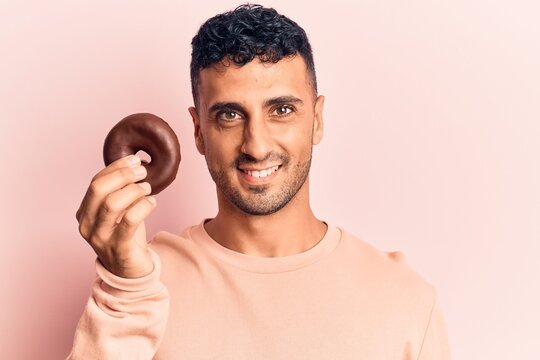Young hispanic man holding donut looking positive and happy standing and smiling with a confident smile showing teeth