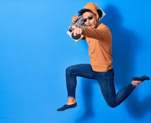 Young handsome latin man smiling happy wearing thug lif sunglasses. Jumping with smile on face holding boombox over isolated blue background