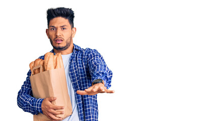 Handsome latin american young man holding paper bag with bread doing stop gesture with hands palms, angry and frustration expression