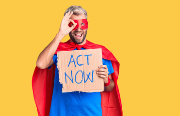 Young blond man wearing super hero costume holding act now cardboard banner smiling happy doing ok...