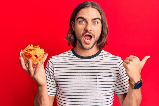Young handsome man holding nachos potato chips pointing thumb up to the side smiling happy with open mouth