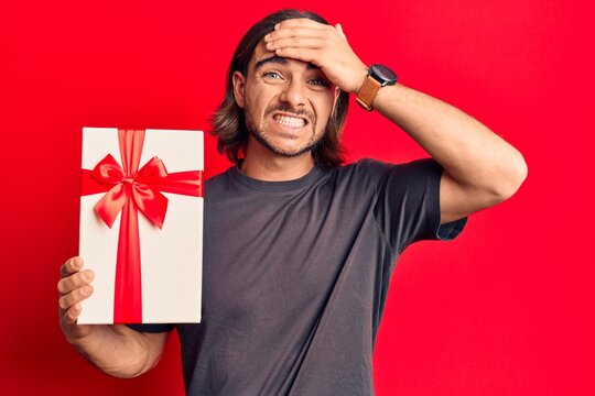 Young handsome man holding gift stressed and frustrated with hand on head, surprised and angry face