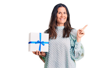 Young beautiful brunette woman holding birthday gift smiling happy pointing with hand and finger to the side