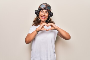 Middle age brunette woman wearing goggles and retro aviator leather hat over white background smiling in love doing heart symbol shape with hands. Romantic concept.