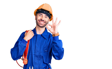 Young hispanic man wearing elecrician uniform holding cable doing ok sign with fingers, smiling friendly gesturing excellent symbol