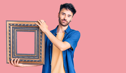 Young hispanic man holding empty frame thinking attitude and sober expression looking self confident