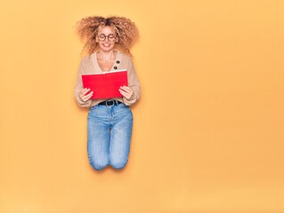 Young beautiful curly student girl wearing glasses smiling happy. Jumping with smile on face reading book over isolated yellow background.