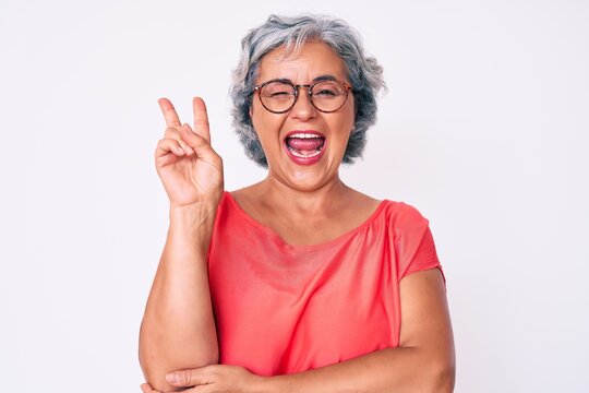 Senior hispanic grey- haired woman wearing casual clothes and glasses smiling with happy face winking at the camera doing victory sign. number two.