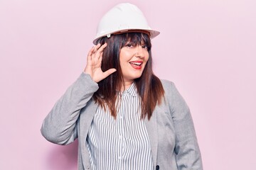 Young plus size woman wearing architect hardhat smiling with hand over ear listening and hearing to rumor or gossip. deafness concept.