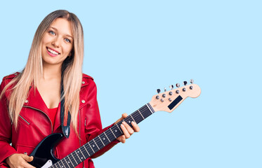 Young beautiful blonde woman playing electric guitar looking positive and happy standing and smiling with a confident smile showing teeth