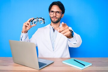 Handsome hispanic optician man at the clinic holding optometry glasses pointing with finger to the camera and to you, confident gesture looking serious