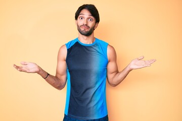 Handsome hispanic man wearing sportswear clueless and confused expression with arms and hands raised. doubt concept.