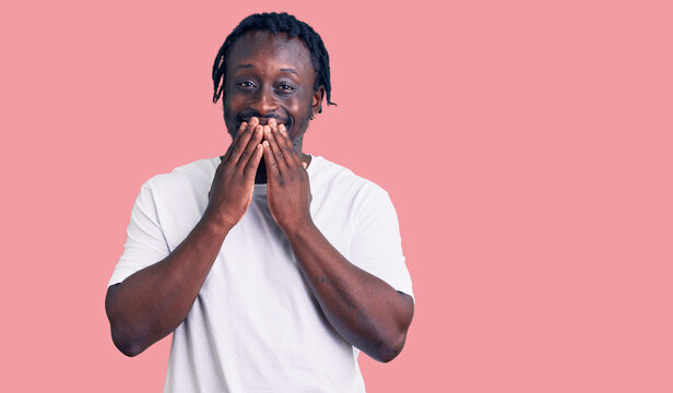 Young african american man with braids wearing casual white tshirt laughing and embarrassed giggle covering mouth with hands, gossip and scandal concept