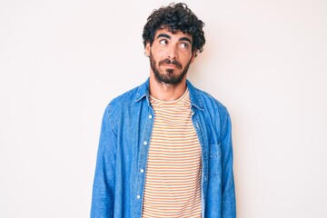 Handsome young man with curly hair and bear wearing casual denim jacket smiling looking to the side and staring away thinking.