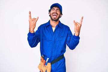 Handsome young man with curly hair and bear weaing handyman uniform shouting with crazy expression doing rock symbol with hands up. music star. heavy music concept.