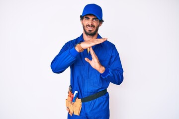 Handsome young man with curly hair and bear weaing handyman uniform doing time out gesture with hands, frustrated and serious face
