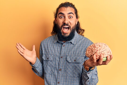 Young arab man holding brain celebrating achievement with happy smile and winner expression with raised hand