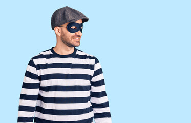 Young handsome man wearing burglar mask looking away to side with smile on face, natural expression. laughing confident.