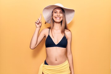 Young beautiful girl wearing bikini and hat smiling with an idea or question pointing finger up with happy face, number one