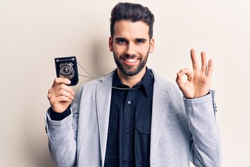 Young handsome man with beard holding police badge doing ok sign with fingers, smiling friendly gesturing excellent symbol