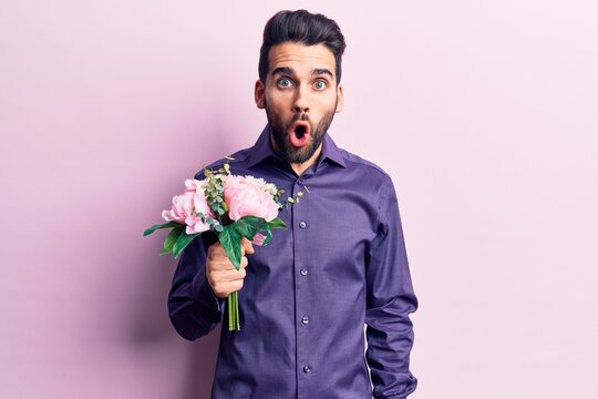 Young handsome man with beard holding bouquet of flowers scared and amazed with open mouth for surprise, disbelief face