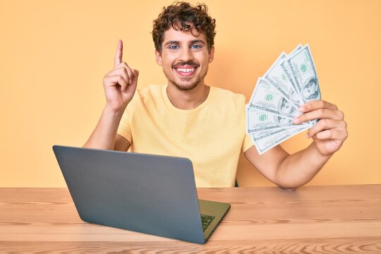Young caucasian man with curly hair working using computer laptop holding usa dollars banknotes smiling with an idea or question pointing finger with happy face, number one