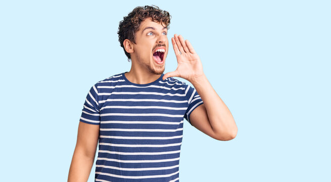 Young handsome man with curly hair wearing casual clothes shouting and screaming loud to side with hand on mouth. communication concept.