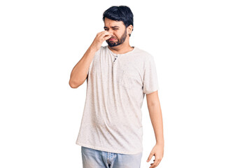 Young hispanic man wearing casual clothes smelling something stinky and disgusting, intolerable smell, holding breath with fingers on nose. bad smell