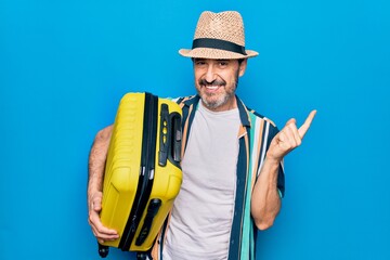 Middle age handsome tourist man on vacation holding cabin bag over isolated blue background smiling happy pointing with hand and finger to the side