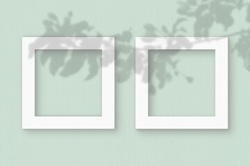 Two square sheet of white textured paper on the light green wall background. Mockup with an overlay of plant shadows. Natural light casts shadows from the leaves of an exotic plant. Flat lay, top view