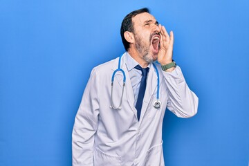 Middle age handsome doctor man wearing coat and stethoscope over isolated blue background shouting and screaming loud to side with hand on mouth. Communication concept.