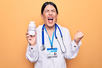 Young doctor woman wearing stethoscope holding bottle of pills over yellow background screaming proud, celebrating victory and success very excited with raised arm