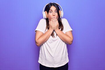 Young beautiful brunette woman listening to music using headphones over purple background covering mouth with hand, shocked and afraid for mistake. Surprised expression