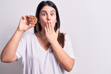 Young beautiful brunette woman eating chocolate cookie over isolated white background covering mouth with hand, shocked and afraid for mistake. Surprised expression