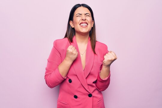 Young beautiful business woman wearing elegant jacket standing over pink background celebrating surprised and amazed for success with arms raised and eyes closed