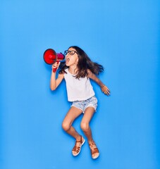 Adorable hispanic child girl wearing glasses. Screaming using megaphone jumping over isolated blue background