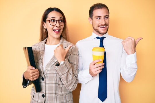 Beautiful couple wearing business clothes drinking take away coffee holding folder pointing thumb up to the side smiling happy with open mouth