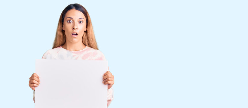 Beautiful young woman holding blank empty banner scared and amazed with open mouth for surprise, disbelief face