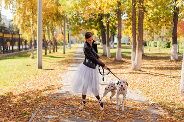 Fototapeta premium Ballerina with Dalmatian dog in the Park. Woman ballerina in a white ballet skirt and black leather jacket dancing in pointe shoes in autumn park with her spotty dalmatian dog.