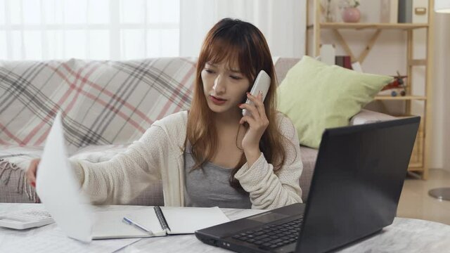 japanese woman is questioning employer on mobile angrily about mistakes in salary causing her high tax. asian female sitting with laptop and document is having phone conversation in anger.