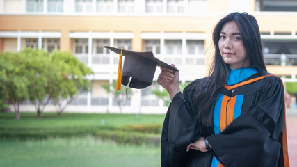 Portrait of happy and excited of young Asian female university graduate wears graduation gown and hat celebrates with degree in university campus in the commencement day. Education concept.