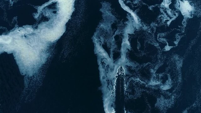 Aerial shot of a fishing boat driving through white run off from a dam creating a visible path through the amazing textures which are like white paint one the blue surface of the water.