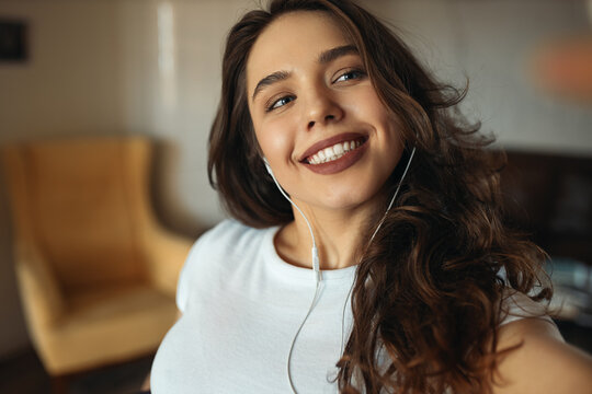 Cropped shot of beautiful young European woman with long curly hair and brown lipstick wearing earphones looking with broad happy smile, taking self portrait using front camera on smart phone