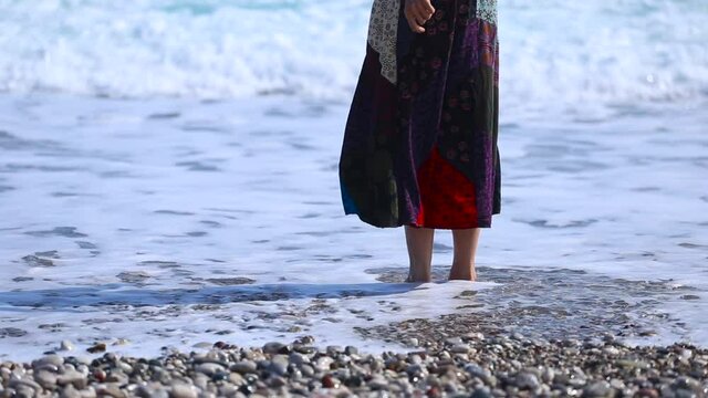 A girl in a long skirt stands on the beach and touches the water with her foot, rest on the seaside, feet in the water, sea waves.