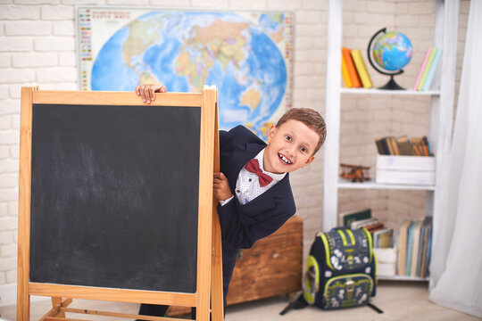 Schoolboy Looks Out From Behind A Black Blackboard In The Classroom.